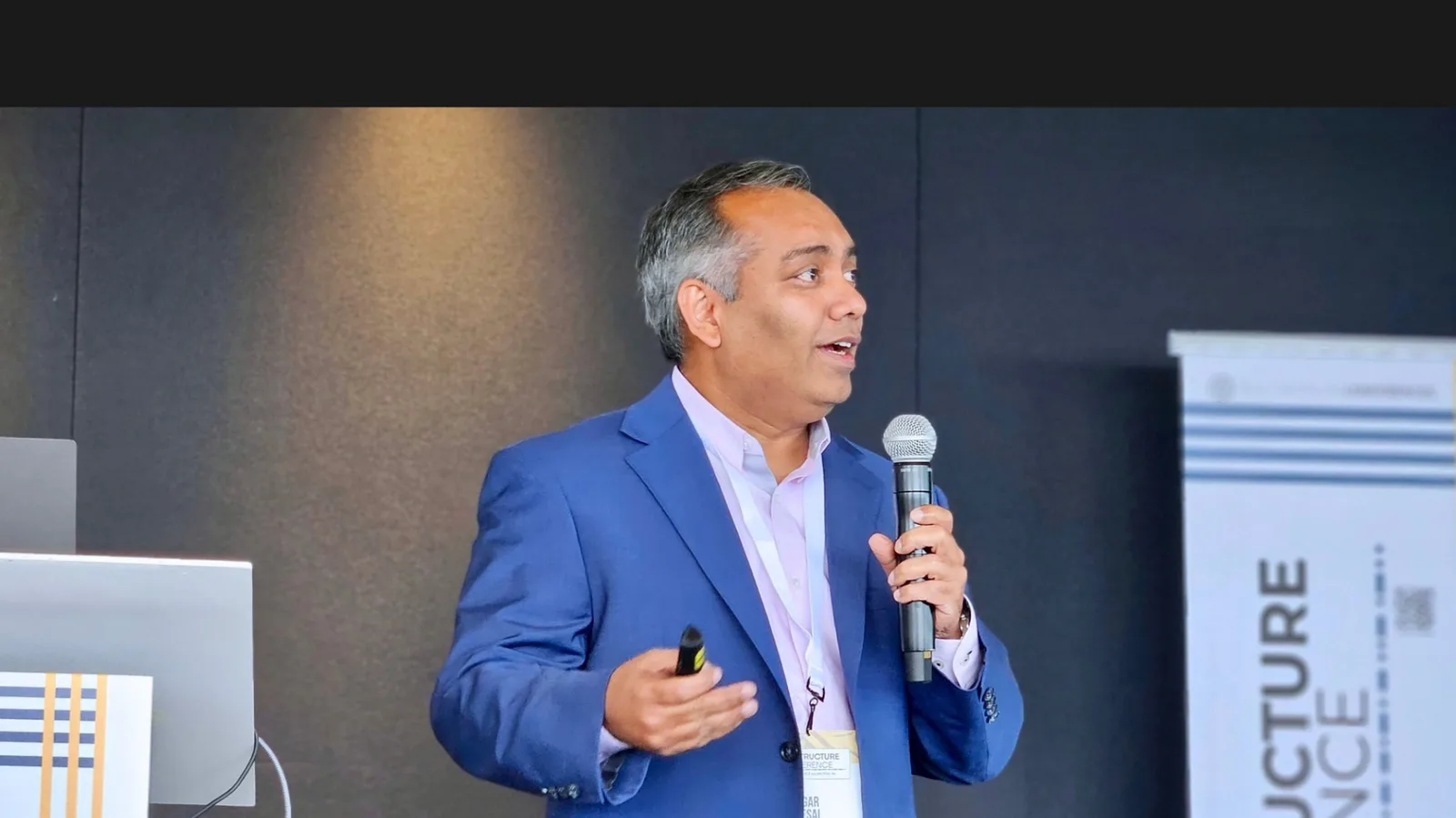 Jigar Desai speaking on stage in blue suit with microphone