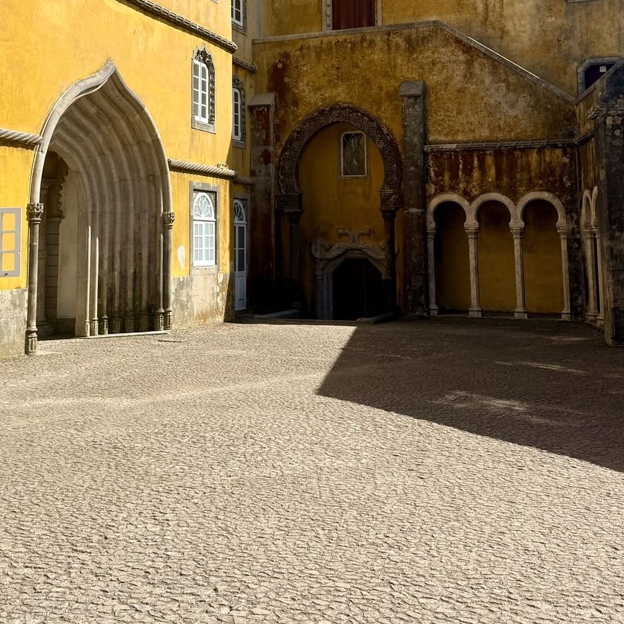 Sunlit courtyard with a Gothic arch, a Moorish arch, and a hard diagonal shadow across the cobblestones, Pena Palace, Sintra, Portugal - 3½ Things August 2026 Edition cover