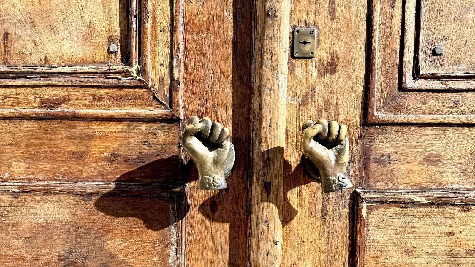 Weathered wooden double door in Lisbon with two cast brass fist knockers throwing hard afternoon shadows